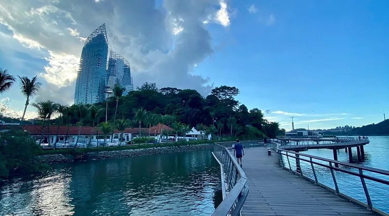 Bukit Chermin Boardwalk over Keppel Bay in late-afternoon light, with a person walking, red-roofed waterfront buildings and the Reflections at Keppel Bay towers above the trees